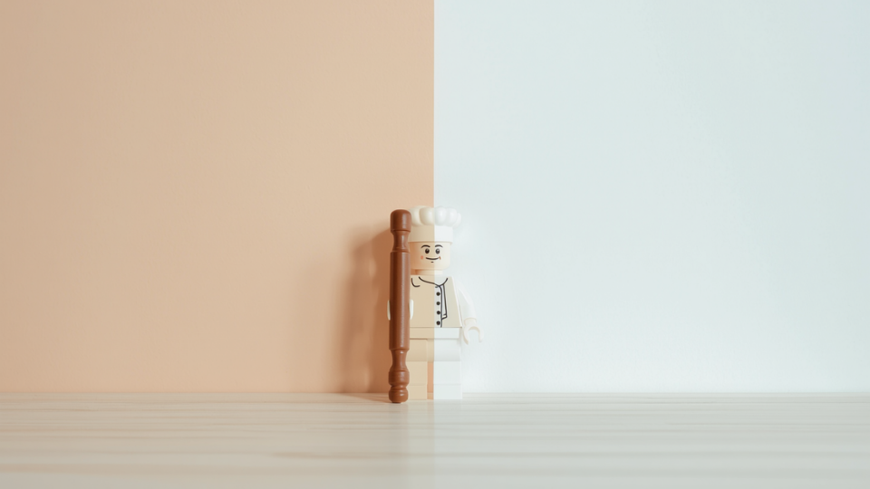 A miniature Lego chef holding a brown rolling pin on a light wooden countertop.