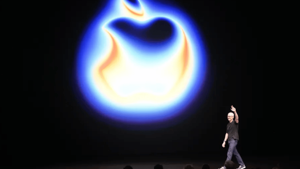 A tech executive stands under a spotlight on a large stage in front of a glowing circular neon logo