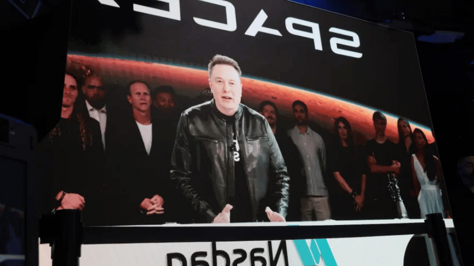 Elon Musk standing on a stage at a corporate event with a giant screen behind him,
