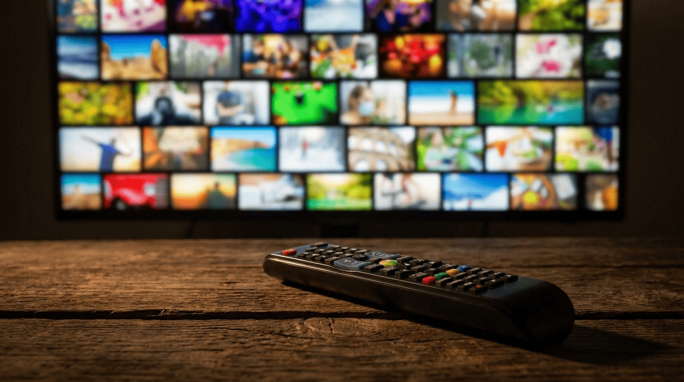 A black television remote control resting on a wooden table in front of a blurry screen showing a grid of video thumbnails.