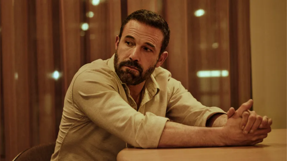 A close-up photograph of actor Ben Affleck sitting at a wooden table in a brightly lit room with soft background lighting.