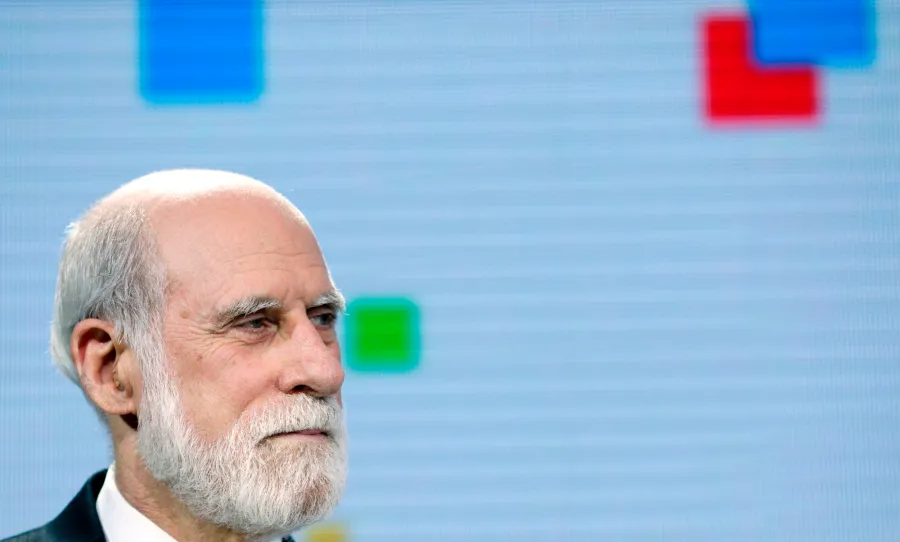 A formal studio portrait of an elderly tech executive with a white beard wearing a dark three-piece suit against a soft blue and green background.
