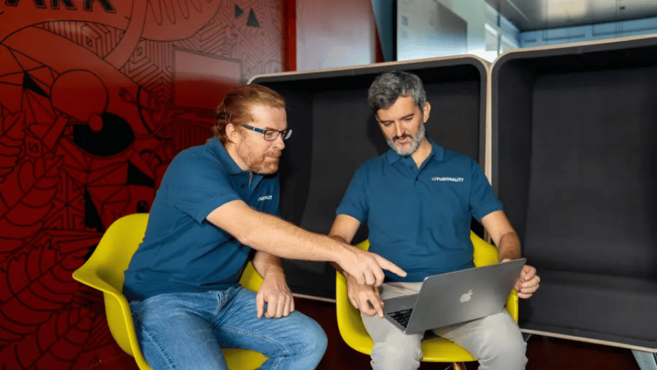 Two engineers sitting in yellow chairs collaborating over a laptop screen.
