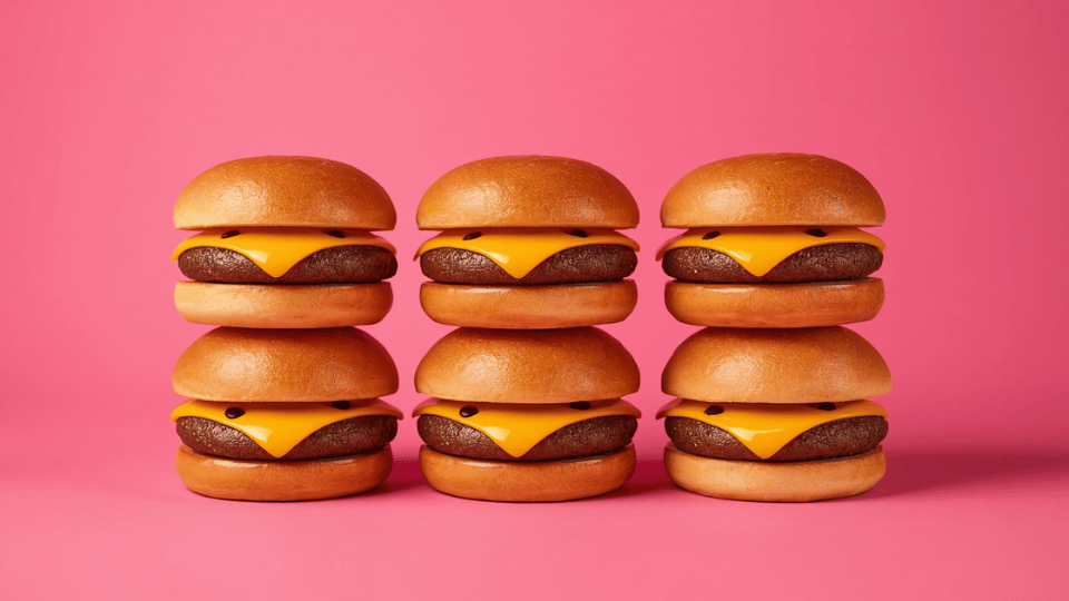 Six identical cheeseburgers lined up neatly against a solid pink background.