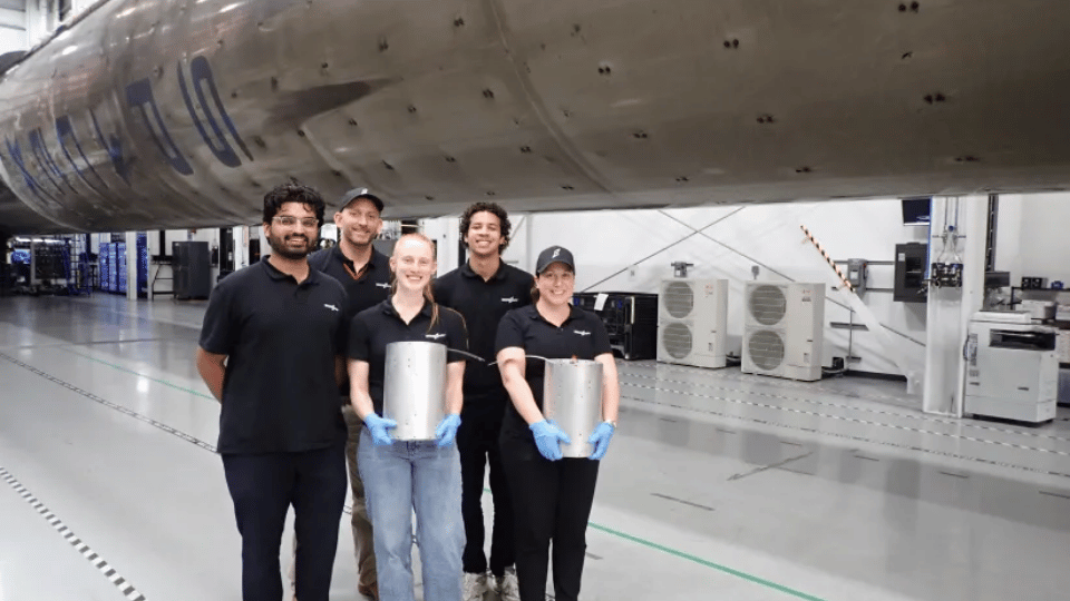 Five engineers posing together inside a hangar in front of a rocket holding metal test canisters.