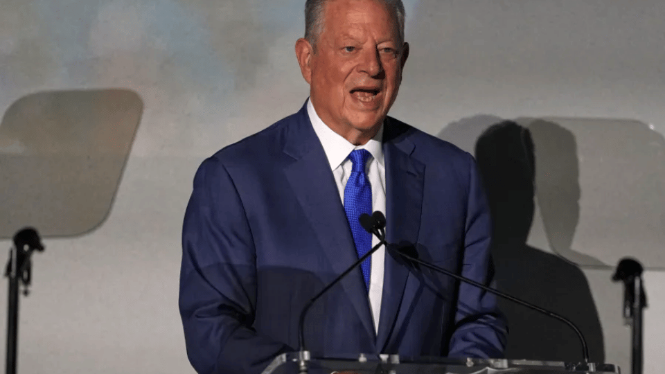 Al Gore standing at a podium wearing a dark blue suit while speaking at a conference.